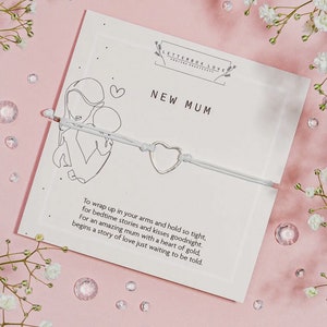 May include: A white card with the text "NEW MUM" and a line drawing of a mother and child. A silver heart-shaped charm is strung on a light gray cord. The card is on a pink surface with flowers and crystals.