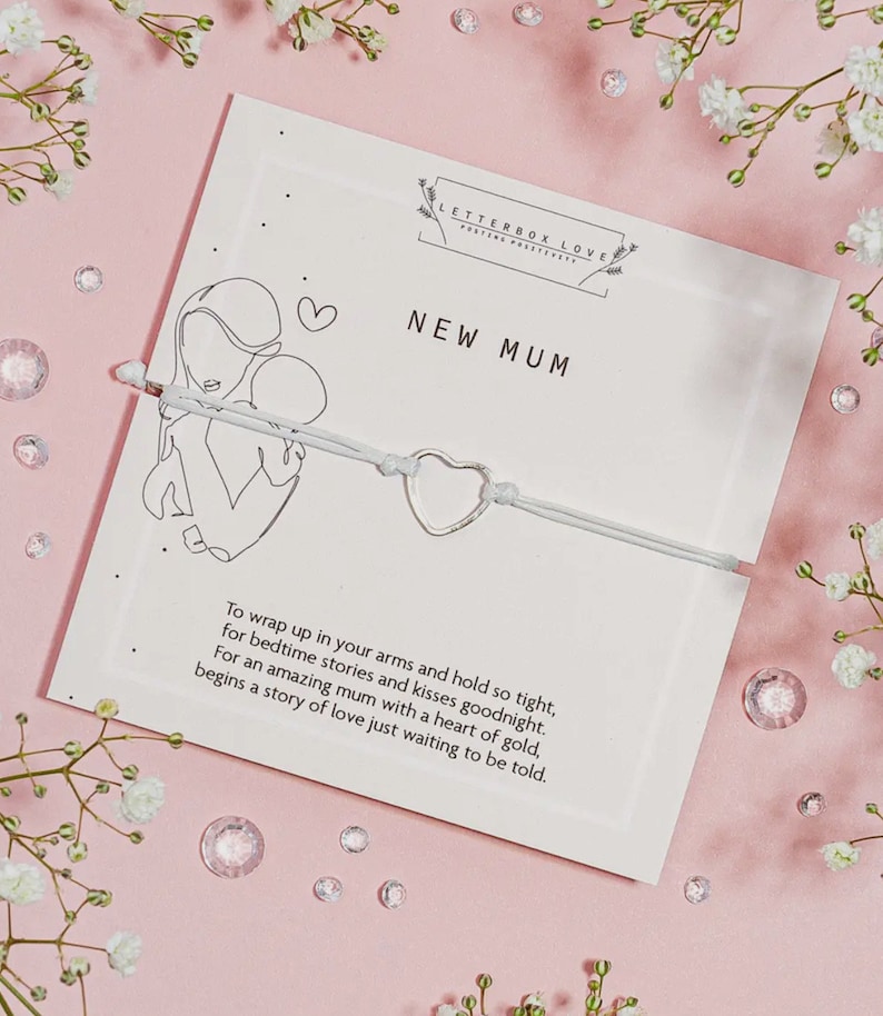 May include: A white card with the text "NEW MUM" and a line drawing of a mother and child. A silver heart-shaped charm is strung on a light gray cord. The card is on a pink surface with flowers and crystals.