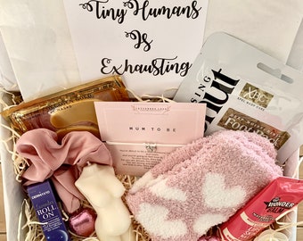 Può includere: Una scatola regalo piena di articoli per una neomamma. La scatola include un calzino di pantofola peloso rosa, una candela bianca a forma di donna incinta, una piccola bottiglia di olio di lavanda roll-on, un elastico per capelli rosa, un biglietto che dice "Mamma a essere" e una maschera per i piedi rosa e bianca.