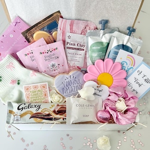 K&ouml;nnte beinhalten: Eine Geschenkbox mit Pflegeprodukten. Enth&auml;lt rosa Gesichtsmasken, Teebeutel, eine Galaxy-Schokoriegel, einen bl&uuml;tenf&ouml;rmigen Keks, einen schwammf&ouml;rmigen Schwamm und eine Karte mit der Aufschrift "A Little Treat for you."