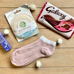 May include: A flat lay of a self-care gift basket with a pink sock, a white facial buffer, a roll-on bottle of lavender essential oil, a bar of Galaxy Cookie Crumble chocolate, and white decorative flowers.
