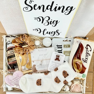 May include: A white gift box filled with various items, including a chocolate bar, a heart-shaped biscuit, a pair of socks with bear faces, a small bear charm, a candle, and a hair scrunchie. The box is decorated with a white card that says "Sending Big Hugs".