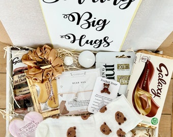 May include: A white gift box filled with various items, including a chocolate bar, a heart-shaped cookie, a pair of socks with bear faces, a small bear charm, a candle, and a hair scrunchie. The box is decorated with a white card that says "Sending Big Hugs".