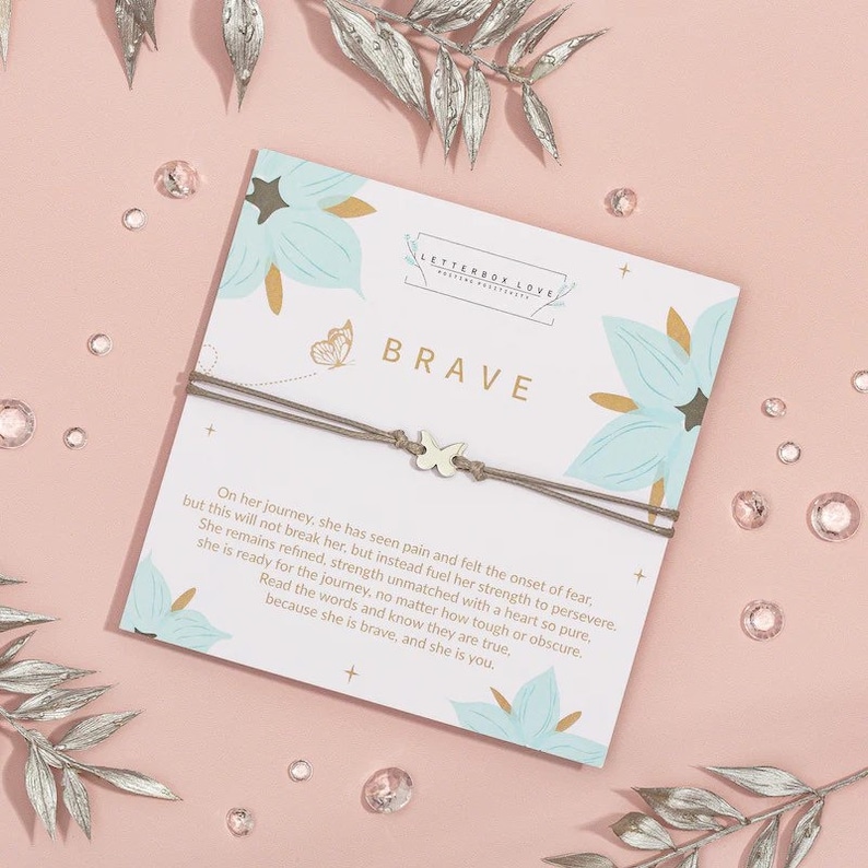 May include: A brown string bracelet with a gold butterfly charm. The bracelet is on a white card with a floral design and the word "BRAVE" in gold lettering. The card also includes a quote about strength and perseverance.