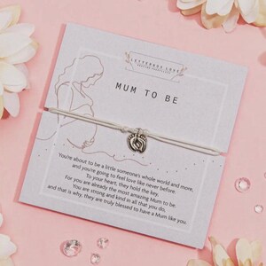 May include: A greeting card with the text "MUM TO BE" and a bracelet with a charm shaped like baby footprints. The card has a drawing of a pregnant person and a message about motherhood. The background is pink with floral accents.