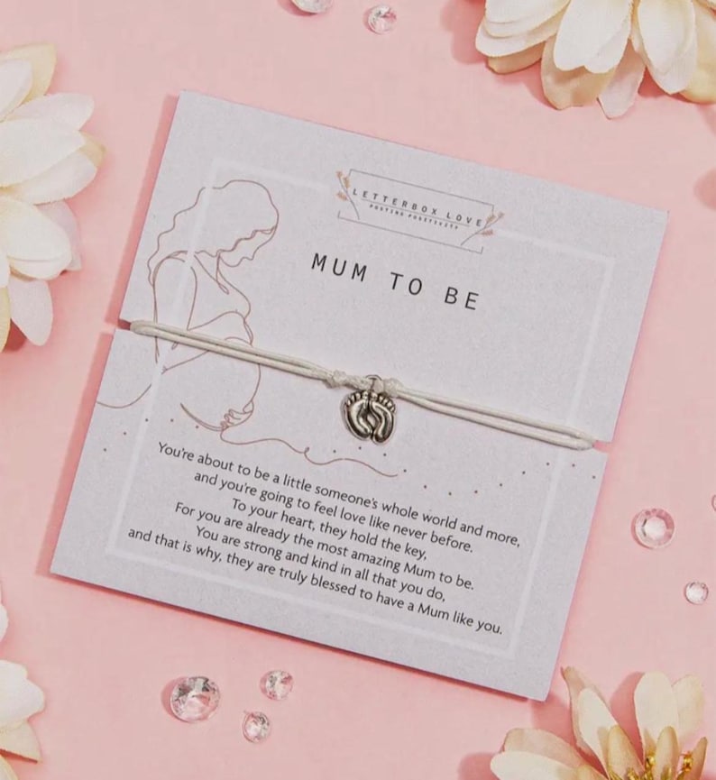 May include: A greeting card with the text "MUM TO BE" and a bracelet with a charm shaped like baby footprints. The card has a drawing of a pregnant person and a message about motherhood. The background is pink with floral accents.