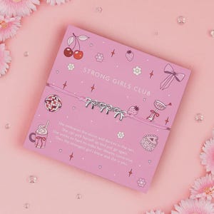 K&ouml;nnte beinhalten: Ein rosa Armband mit silbernen Schleifenanh&auml;ngern wird auf einer rosa Karte mit dem Text "STRONG GIRLS CLUB" pr&auml;sentiert. Die Karte zeigt Illustrationen von Kirschen, Erdbeeren und anderen dekorativen Elementen. Das Armband befindet sich auf einem rosa Hintergrund.