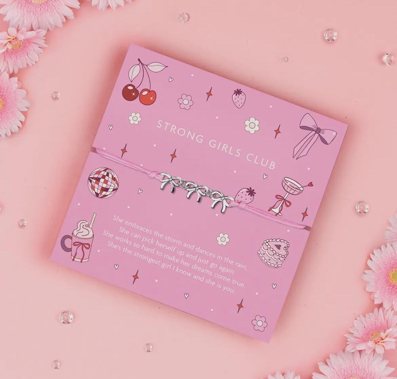 K&ouml;nnte beinhalten: Ein rosa Armband mit silbernen Schleifenanh&auml;ngern wird auf einer rosa Karte mit dem Text "STRONG GIRLS CLUB" pr&auml;sentiert. Die Karte zeigt Illustrationen von Kirschen, Erdbeeren und anderen dekorativen Elementen. Das Armband befindet sich auf einem rosa Hintergrund.