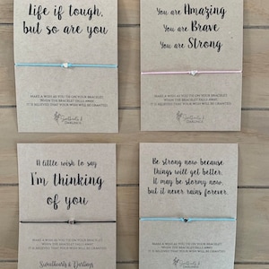 May include: Four kraft paper cards with inspirational messages and blue and pink string bracelets. The cards have the text "Life if tough, but so are you", "You are Amazing, You are Brave, You are Strong", "A little wish to say I'm thinking of you", and "Be strong now because things will get better. It may be stormy now, but it never rains forever". Each card has a small silver charm on the string bracelet.
