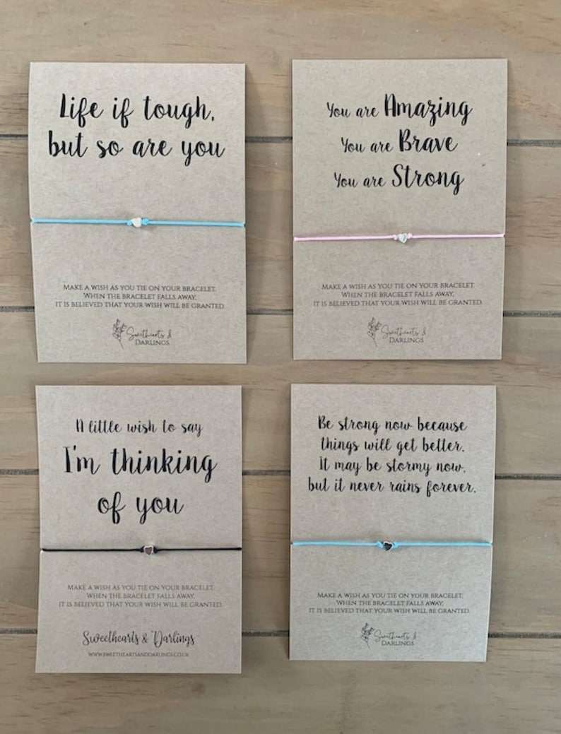 May include: Four kraft paper cards with inspirational messages and blue and pink string bracelets. The cards have the text "Life if tough, but so are you", "You are Amazing, You are Brave, You are Strong", "A little wish to say I'm thinking of you", and "Be strong now because things will get better. It may be stormy now, but it never rains forever". Each card has a small silver charm on the string bracelet.