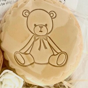 May include: A round, beige cookie with a teddy bear design in the center. The bear is outlined in a darker shade of brown, with a bow tie. The cookie has a scalloped edge and is packaged in clear cellophane.