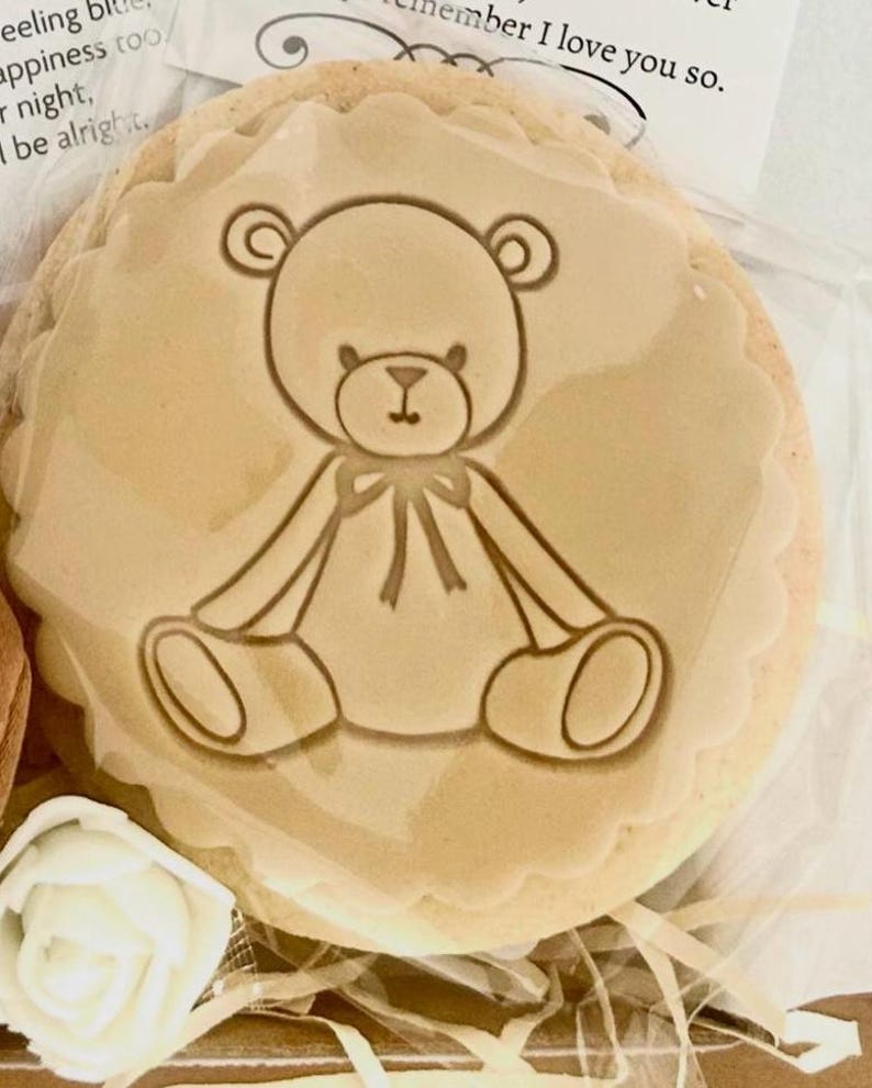 May include: A round, beige cookie with a teddy bear design in the center. The bear is outlined in a darker shade of brown, with a bow tie. The cookie has a scalloped edge and is packaged in clear cellophane.