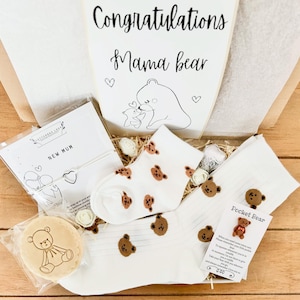 May include: A gift box with a "Congratulations Mama Bear" card, white socks with brown bear faces, a "New Mum" card, and a bear-shaped cookie. The box includes decorative flowers and a "Pocket Bear" card.