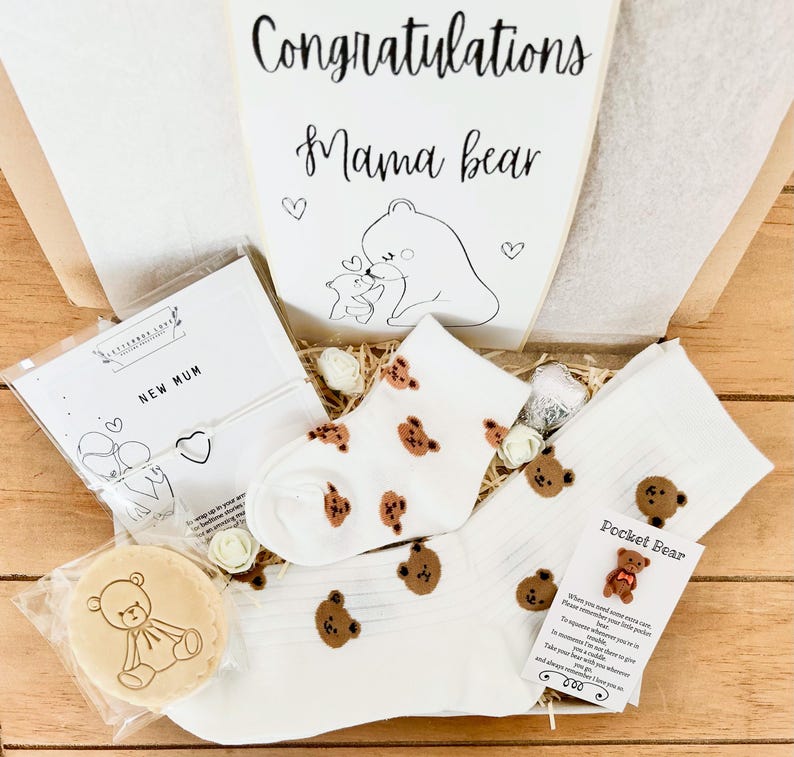 May include: A gift box with a "Congratulations Mama Bear" card, white socks with brown bear faces, a "New Mum" card, and a bear-shaped cookie. The box includes decorative flowers and a "Pocket Bear" card.