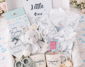 May include: A white gift box containing a Welcome Little One card, a plush gray elephant, two white mugs labeled Daddy's Mug and Mummy's Mug, gray knit baby booties, a baby outfit with elephant prints, a Hello Baby milestone card set, and a heart sign.