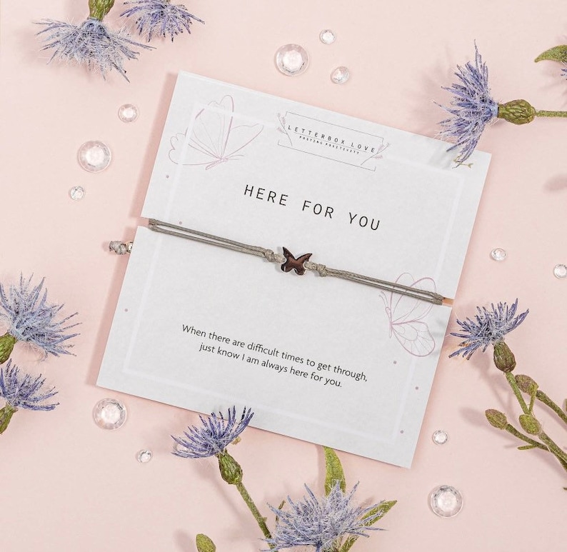May include: A gray string bracelet with a small butterfly charm. The bracelet is on a white card with the text "HERE FOR YOU" and a message about being there for someone during difficult times.
