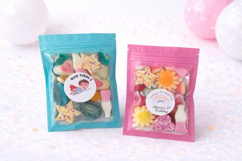 K&ouml;nnte beinhalten: Zwei wiederverschlie&szlig;bare Beutel gef&uuml;llt mit gemischten Fruchtgummis. Der t&uuml;rkisfarbene Beutel hat einen Aufkleber mit der Aufschrift Ollie Turns 3 und einem Superhelden. Der rosa Beutel zeigt den Text Thank you for celebrating Eleonor's 6th Birthday.