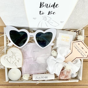 May include: A bridal gift box with the text "Bride to Be" contains heart-shaped sunglasses, a cookie, a candle, and other accessories. The box includes white and gold items, ideal for a bride-to-be.