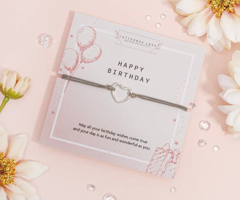 K&ouml;nnte beinhalten: Ein graues Armband aus Schnur mit einem silbernen Herz-Anh&auml;nger und einer Karte mit der Aufschrift "Happy Birthday" mit rosa und wei&szlig;en Ballons und Sternen. Die Karte sagt auch "M&ouml;gen all deine Geburtstagsw&uuml;nsche in Erf&uuml;llung gehen und dein Tag so lustig und wunderbar sein wie du."