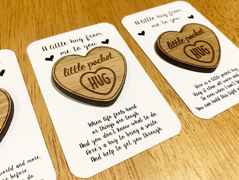 May include: A wooden heart-shaped keychain with the text "little pocket hug" engraved on it. The keychain is attached to a white card with a message about hugs and how they can help when life is tough.