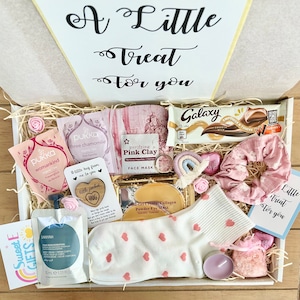 May include: A gift box filled with self-care items. Includes a Galaxy chocolate bar, face mask, tea bags, a candle, a pair of socks with pink hearts, a scrunchie, and a small bag of bath salts. The box has the text "A Little Treat For You".