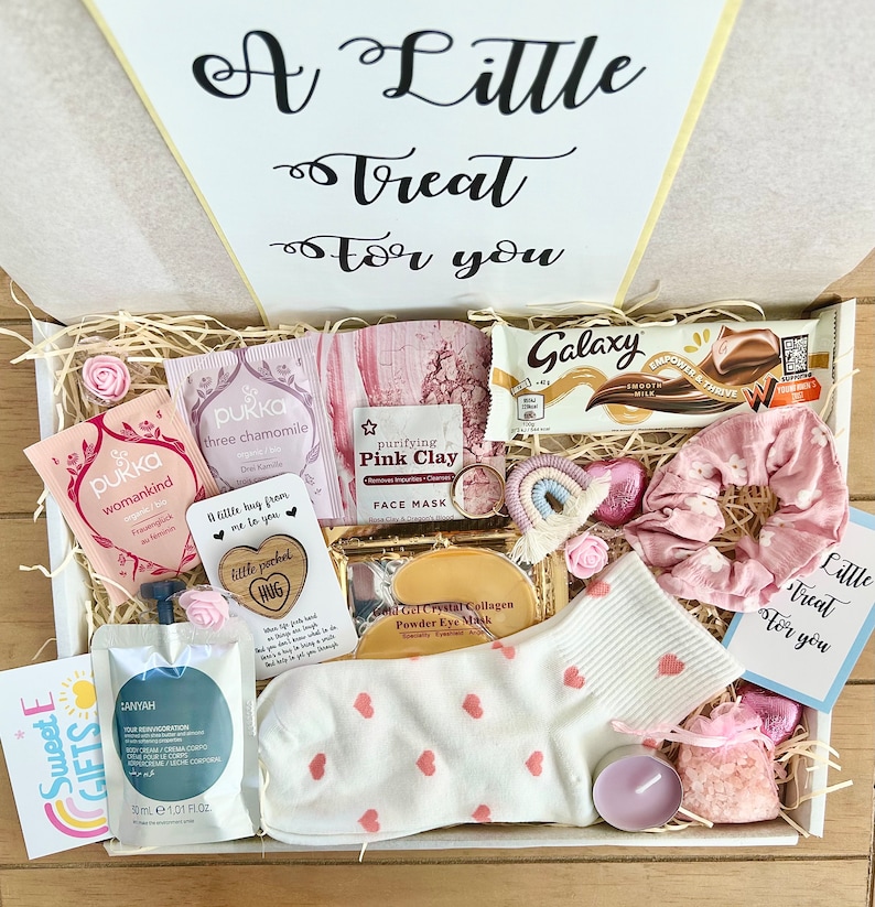 May include: A gift box filled with self-care items. Includes a Galaxy chocolate bar, face mask, tea bags, a candle, a pair of socks with pink hearts, a scrunchie, and a small bag of bath salts. The box has the text "A Little Treat For You".