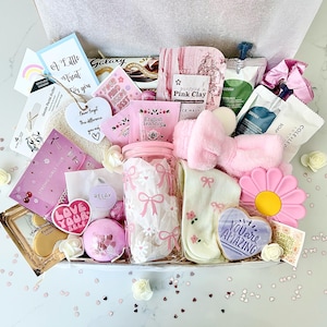 K&ouml;nnte beinhalten: Eine Geschenkbox mit Selbstpflegeartikeln in Rosa, darunter ein Glas mit Schleifen, Gesichtsmasken, ein Stirnband, Socken und ein herzf&ouml;rmiger Keks mit der Aufschrift "You are Amazing".