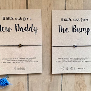 Puede incluir: Dos tarjetas con pulseras de deseos adjuntas. La tarjeta de la izquierda dice "Un peque&ntilde;o deseo para un nuevo pap&aacute;". La tarjeta de la derecha dice "Un peque&ntilde;o deseo de la protuberancia". Ambas tarjetas tienen instrucciones para hacer un deseo al atar la pulsera y que cuando la pulsera se caiga, se cree que tu deseo se conceder&aacute;.