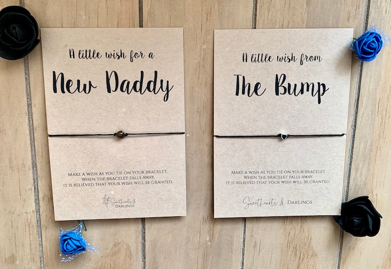 Puede incluir: Dos tarjetas con pulseras de deseos adjuntas. La tarjeta de la izquierda dice "Un peque&ntilde;o deseo para un nuevo pap&aacute;". La tarjeta de la derecha dice "Un peque&ntilde;o deseo de la protuberancia". Ambas tarjetas tienen instrucciones para hacer un deseo al atar la pulsera y que cuando la pulsera se caiga, se cree que tu deseo se conceder&aacute;.