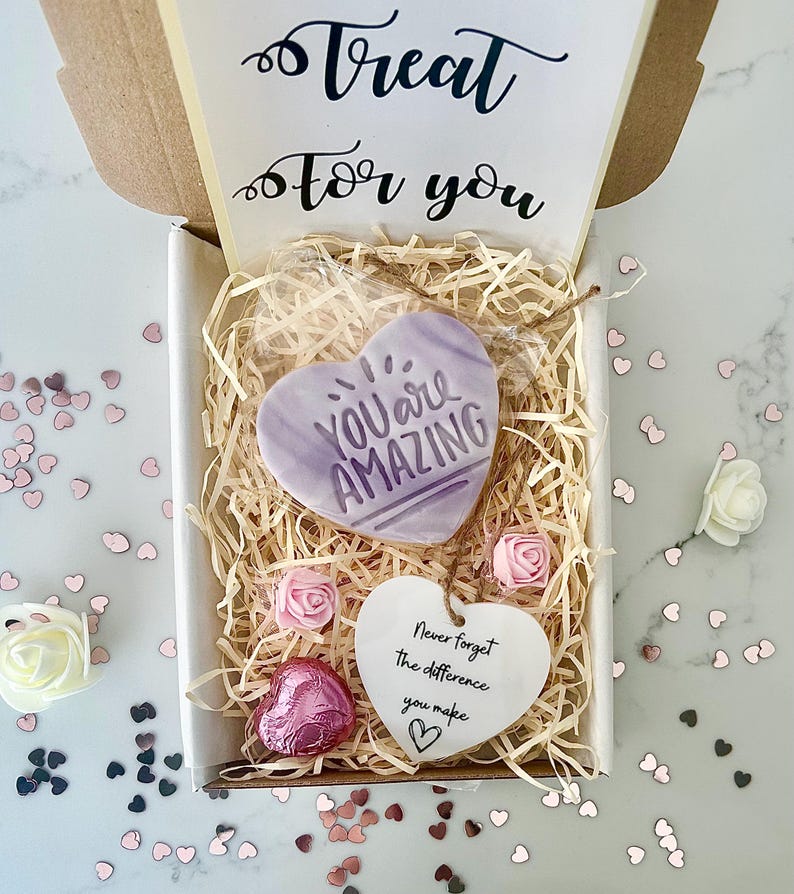 K&ouml;nnte beinhalten: Eine Geschenkbox mit einem herzf&ouml;rmigen Leckerli mit der Aufschrift "You Are Amazing". Die Box enth&auml;lt Dekopapier, rosa Rosen, eine herzf&ouml;rmige Schokolade und ein Herz-Tag mit der Aufschrift "Never forget the difference you make."