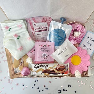 K&ouml;nnte beinhalten: Eine Geschenkbox mit verschiedenen Pflegeprodukten. Sie enth&auml;lt wei&szlig;e Socken mit rosa Schleifen, eine rosa Gesichtsmaske, Teebeutel, Seife, einen Schokoriegel, eine rosa bl&uuml;tenf&ouml;rmige Seife und eine Karte mit dem Text "A Little Treat For You".