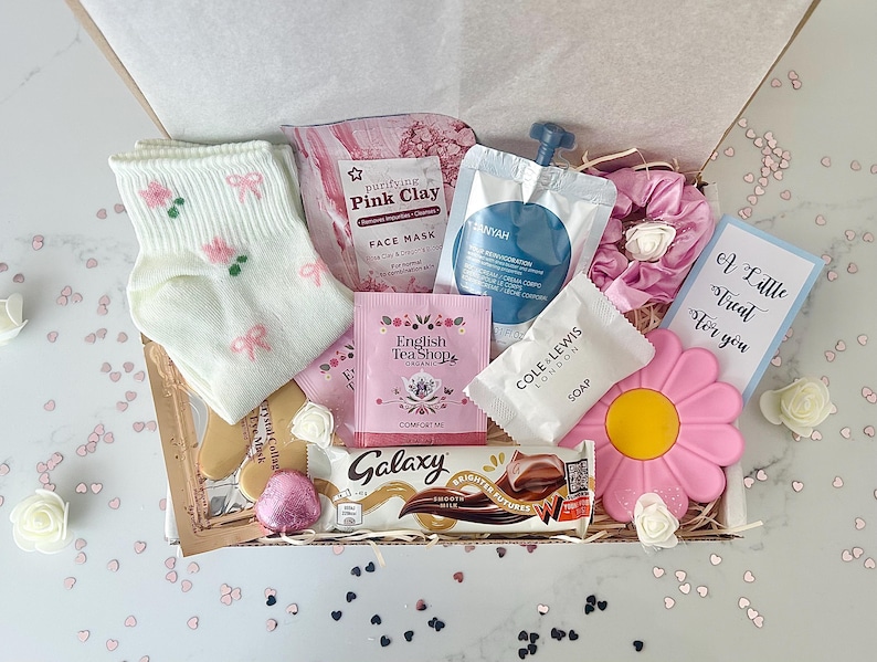 K&ouml;nnte beinhalten: Eine Geschenkbox mit verschiedenen Pflegeprodukten. Sie enth&auml;lt wei&szlig;e Socken mit rosa Schleifen, eine rosa Gesichtsmaske, Teebeutel, Seife, einen Schokoriegel, eine rosa bl&uuml;tenf&ouml;rmige Seife und eine Karte mit dem Text "A Little Treat For You".
