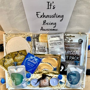 Puede incluir: Una caja de regalo blanca llena de art&iacute;culos de cuidado personal, que incluyen mascarillas faciales, mascarillas para pies, mascarillas para ojos, productos de ba&ntilde;o y velas. La caja tiene una tapa blanca con el texto "It's Exhausting Being Awesome" escrito en negro.