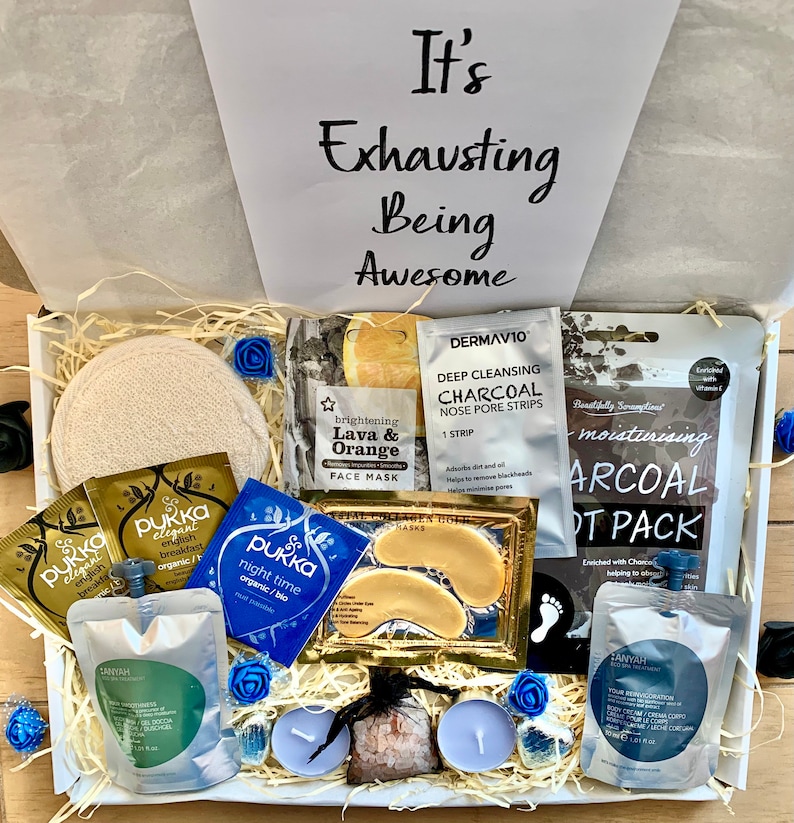 Puede incluir: Una caja de regalo blanca llena de art&iacute;culos de cuidado personal, que incluyen mascarillas faciales, mascarillas para pies, mascarillas para ojos, productos de ba&ntilde;o y velas. La caja tiene una tapa blanca con el texto "It's Exhausting Being Awesome" escrito en negro.