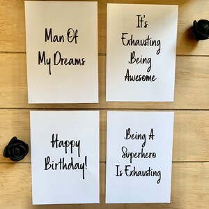 Puede incluir: Cuatro tarjetas blancas con texto escrito a mano en negro. Las tarjetas dicen "Man Of My Dreams", "It's Exhausting Being Awesome", "Happy Birthday!" y "Being A Superhero Is Exhausting".