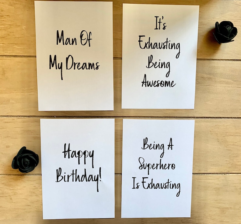 Puede incluir: Cuatro tarjetas blancas con texto escrito a mano en negro. Las tarjetas dicen "Man Of My Dreams", "It's Exhausting Being Awesome", "Happy Birthday!" y "Being A Superhero Is Exhausting".