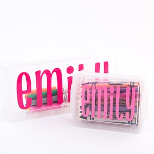 May include: Two clear plastic storage boxes with the name "emily" in bright pink lettering. One box contains markers, the other contains crayons. The boxes are on a white background.