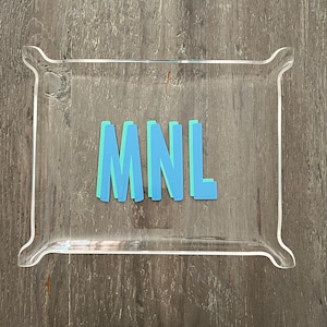 May include: A clear acrylic tray with rounded edges and a raised lip. The tray has the letters "MNL" written in blue and green on the inside.