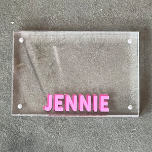 May include: A clear acrylic picture frame with a pink inscription that reads "JENNIE".