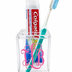 May include: A clear acrylic toothbrush holder featuring a pink monogram design on the front. Inside the holder is a tube of Colgate toothpaste with a net weight of 4.8 ounces and a white toothbrush with blue bristles and a teal handle.