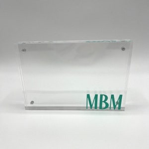 May include: A clear acrylic picture frame with a teal green inscription that reads "MBM".