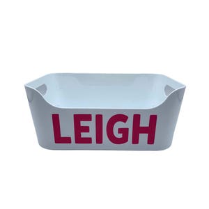 May include: White plastic storage bin with a handle on each side. The bin has the name "LEIGH" printed in pink letters.