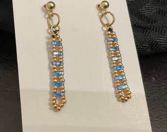 May include: A pair of gold dangle earrings with blue and gold beads. The earrings are on a white card.