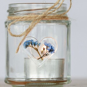 Pode incluir: Um frasco de vidro transparente com um design de flores prensadas em forma de coração e uma vela de chá acesa dentro. O frasco é amarrado com um barbante.