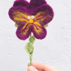 May include: A purple felt flower with a yellow butterfly design and green felt leaves on a stem.