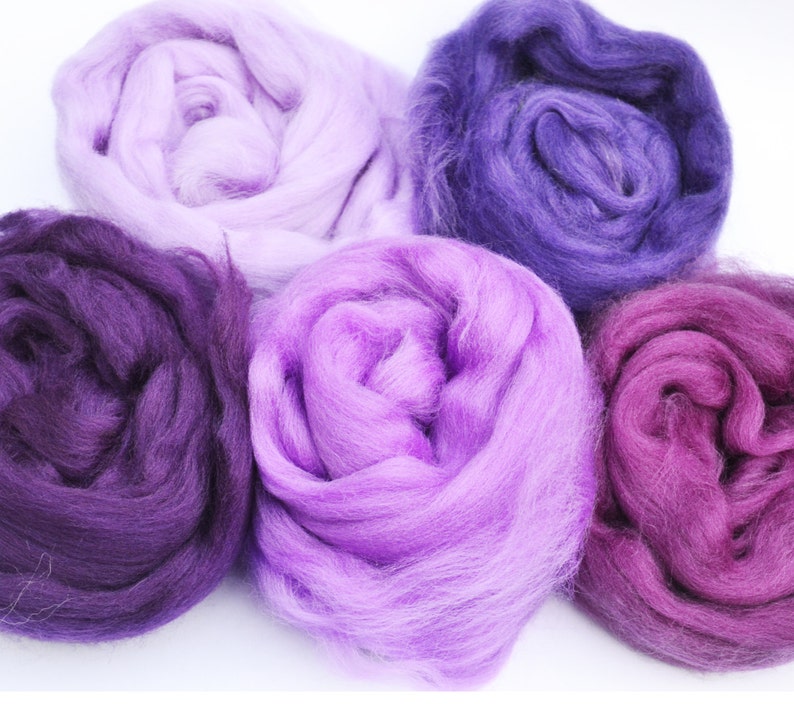 Puede incluir: Cinco ovillos de lana en tonos de morado, lavanda y rosa. La lana es suave y esponjosa, perfecta para tejer o hacer ganchillo.