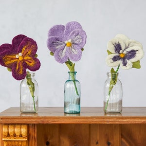 May include: Three felt flower arrangements in glass vases. The flowers are purple, white, and a deep purple with yellow accents. The vases are clear glass with a textured surface. The flowers are sitting on a wooden shelf.