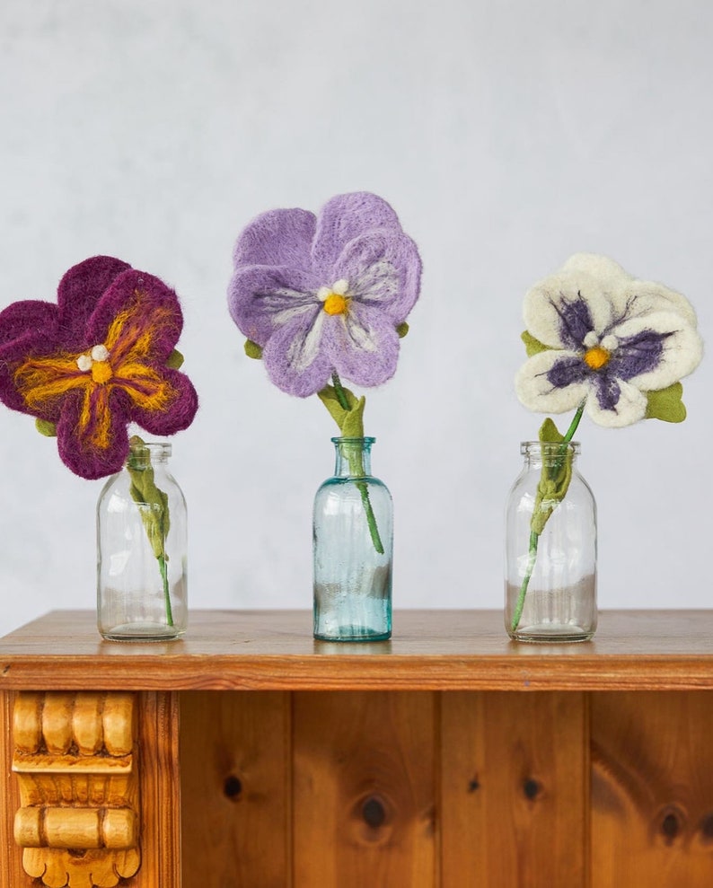 May include: Three felt flower arrangements in glass vases. The flowers are purple, white, and a deep purple with yellow accents. The vases are clear glass with a textured surface. The flowers are sitting on a wooden shelf.