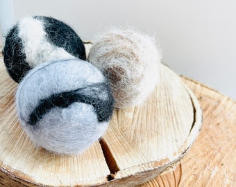 May include: Three felted wool balls in black and white, gray and black, and beige. The balls are sitting on a wooden surface.
