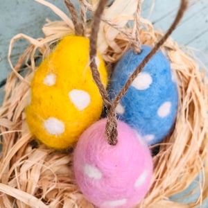 May include: Three colorful Easter egg ornaments in a nest of straw. The eggs are yellow, blue, and pink, each with white polka dots. They are suspended by twine loops, perfect for hanging. A festive and decorative item.